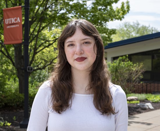 Bella Stephens, in white top with brown hair down past shoulders, stands in front of foliage on campus.